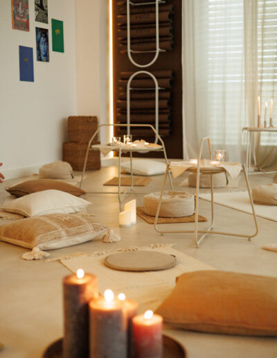 Ein gemütlicher Meditationsraum mit Kissen, Matten und brennenden Kerzen. Weiches natürliches Licht fällt durch die Vorhänge. Yogamatten liegen aufgerollt an einem Wandregal, perfekt für Pilates und Yoga in Bruchsal. Kunst schmückt die weißen Wände und schafft eine ruhige Atmosphäre.
