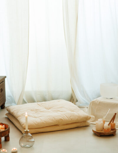 Ein gemütlicher Meditationsraum mit Bodenkissen, Kerzen, einem Holztablett mit Kristallen und Klangschalen vor weißen Vorhängen - perfekt für Coaching oder Pilates und Yoga in Bruchsal, um eine ruhige und gelassene Atmosphäre zu schaffen.