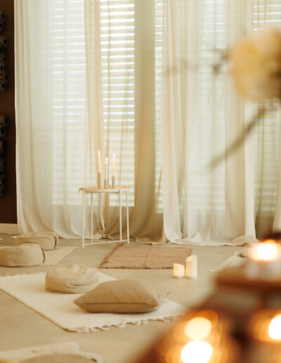 Ein sanft beleuchteter Raum mit durchsichtigen Vorhängen, Yogamatten, Kissen, kleinen Tischen und brennenden Kerzen schafft eine ruhige und gemütliche Atmosphäre - perfekt für Pilates und Yoga in Bruchsal oder Coaching-Sitzungen. Aufgerollte Matten stapeln sich an der Wand, Blumen verschwimmen im Vordergrund.
