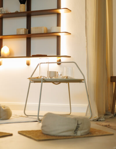 Ein ruhiger, minimalistischer Raum für Pilates und Yoga in Bruchsal, mit Bodenkissen, niedrigen Matten, einem weißen Loungesessel und Holzregalen mit Dekoration. Sanfte Beleuchtung und eine ruhige Einrichtung bieten einen friedlichen Raum, der ideal für Entspannung und Coaching ist.
