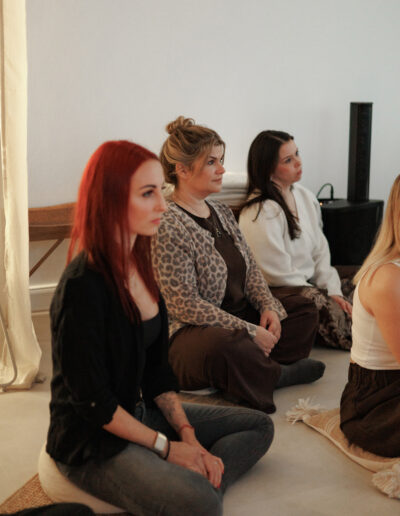 Drei Frauen sitzen im Schneidersitz auf Matten in einem Raum, schauen nach vorne und wirken aufmerksam. Sie sind leger gekleidet in einem minimalistischen Raum mit neutralen Tönen - ein idealer Rahmen für Coaching, Pilates und Yoga in Bruchsal.