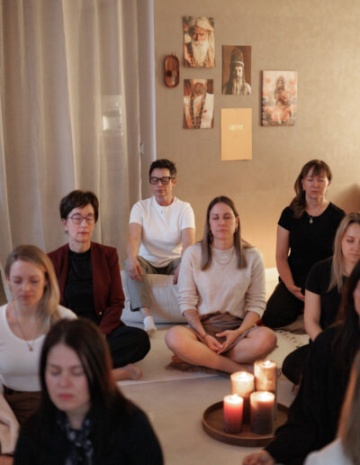 Eine Gruppe von Menschen sitzt im Schneidersitz auf dem Boden in einem ruhigen Raum bei Coaching, Pilates und Yoga in Bruchsal. Die Augen in Meditation geschlossen, umgeben von brennenden Kerzen und spirituellen Kunstwerken, die eine friedliche Atmosphäre schaffen.