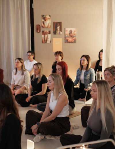 Eine Gruppe von Menschen sitzt im Schneidersitz auf Kissen in einem sanft beleuchteten Raum, die Augen geschlossen, und meditiert. Die friedliche Atmosphäre deutet auf ganzheitliche Praktiken wie Pilates und Yoga in Bruchsal oder Coaching hin, mit beruhigender Kunst und weißen Vorhängen im Hintergrund.