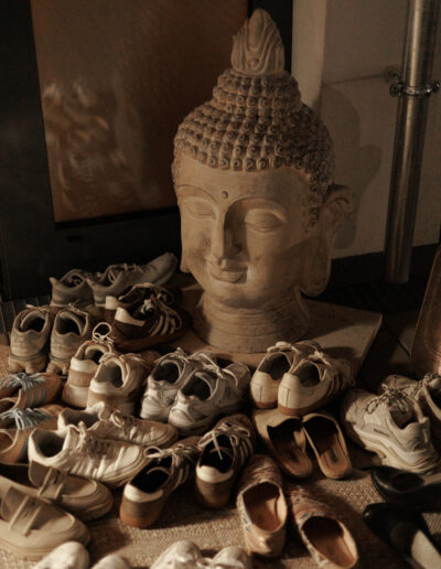 Eine große Buddha-Kopf-Statue ist umgeben von Schuhen und Turnschuhen und schafft einen friedlichen und doch belebten Eingangsbereich - eine einladende Szene, die an ein Pilates und Yoga in Bruchsal Studio oder einen Coachingraum erinnert.
